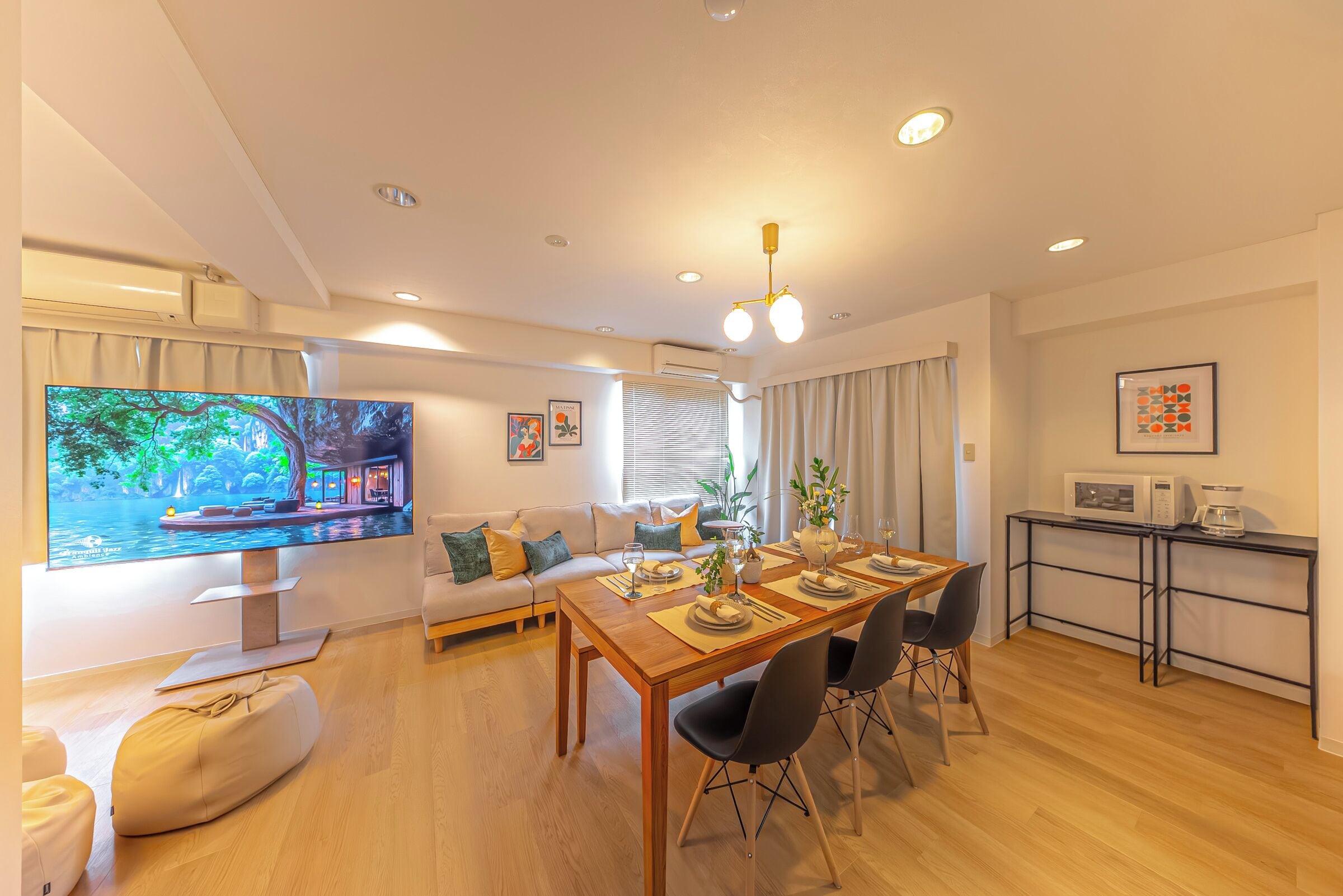 A modern living room with a wooden dining table, black chairs, a sofa, a flat-screen TV, and a kitchen area with a microwave and coffee maker.