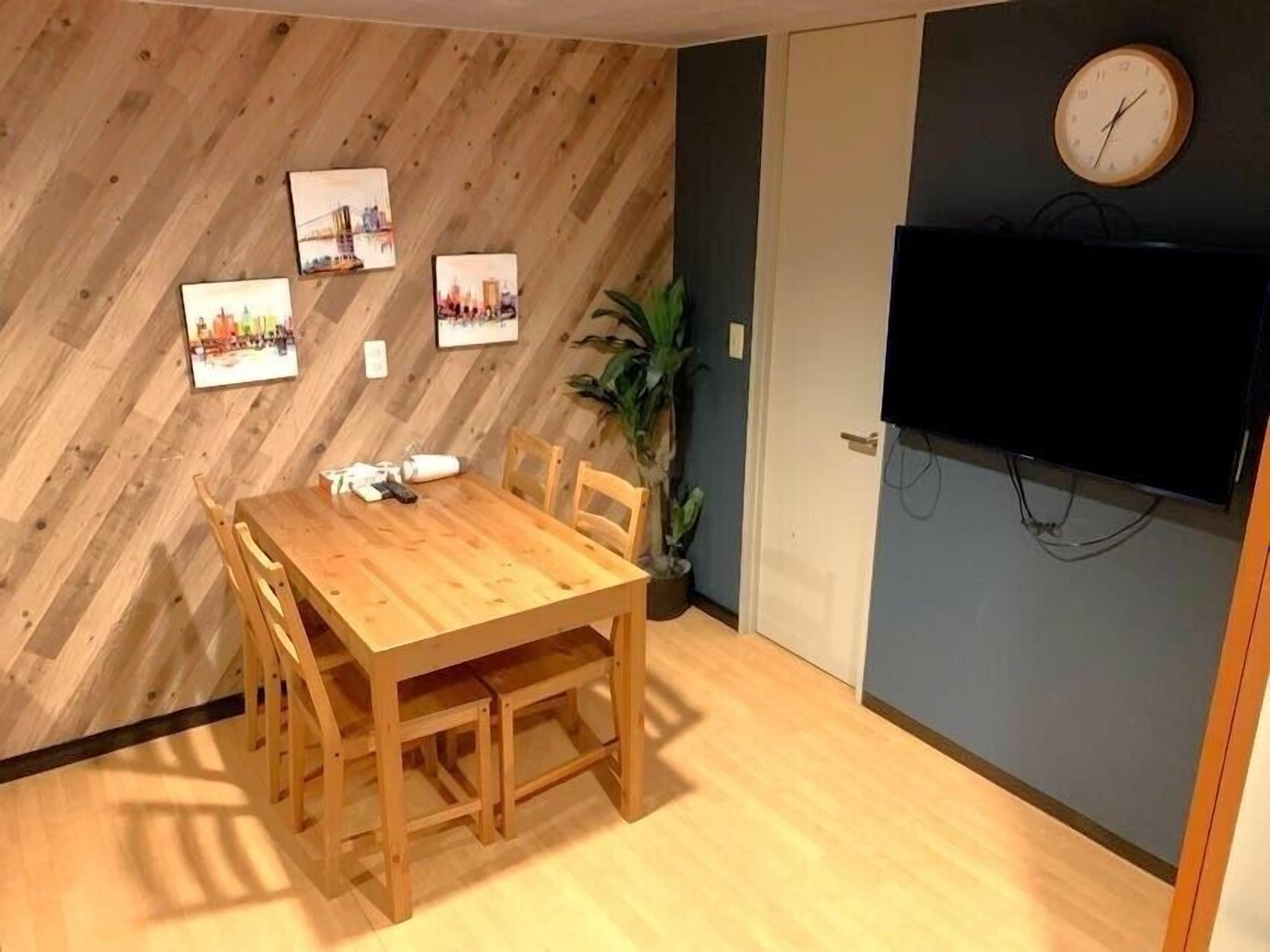A dining area with a wooden table and chairs, a clock on the wall, and a flat-screen TV mounted above the table.