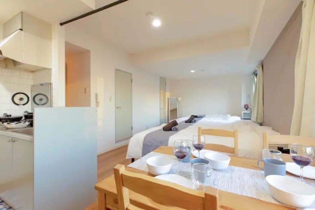 A modern hotel room with a bed, a dining table set with wine glasses and bowls, and a kitchenette with a refrigerator and microwave.