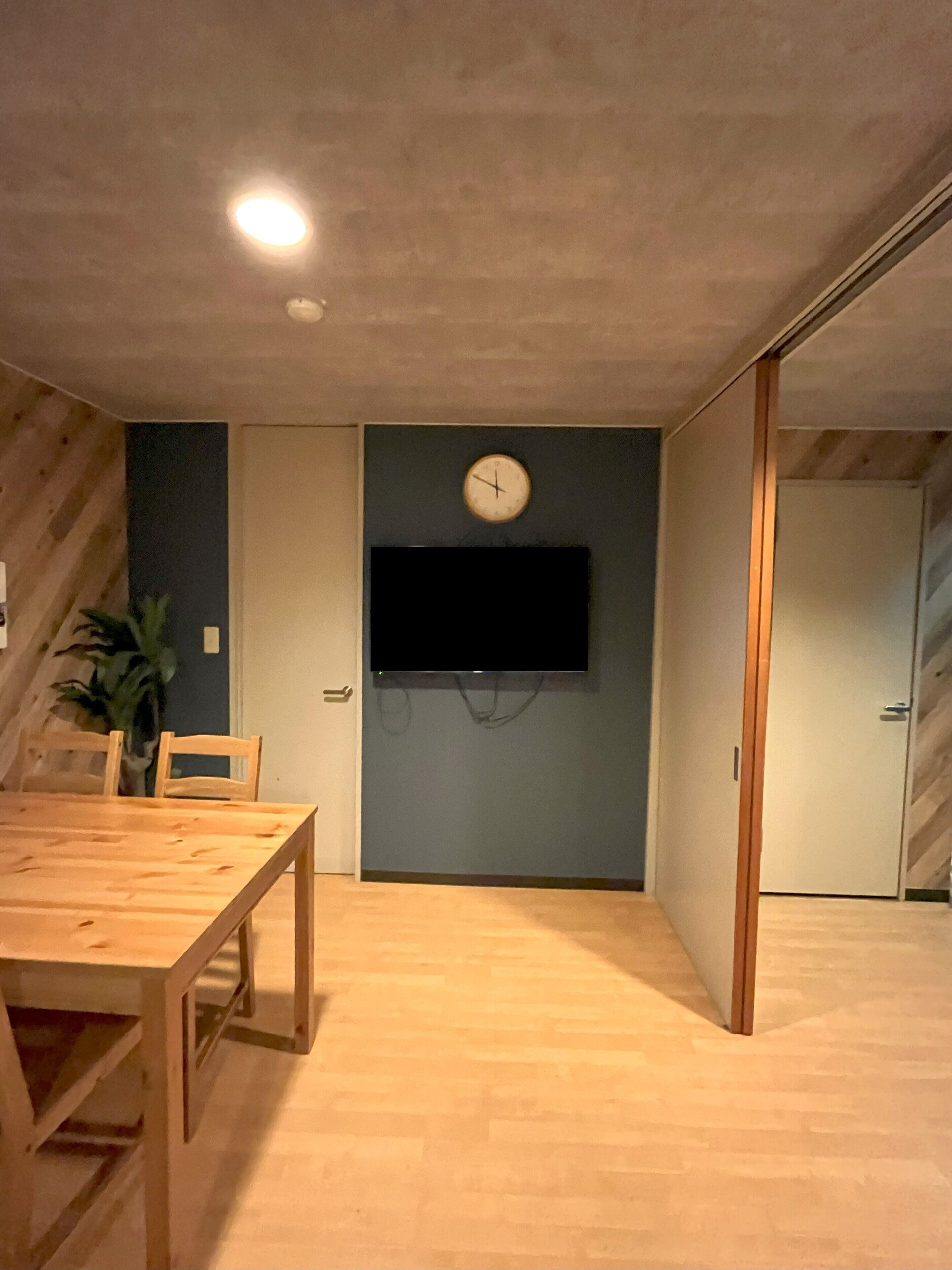A room with a wooden dining table, chairs, a wall-mounted clock, a flat-screen TV, and a sliding door.