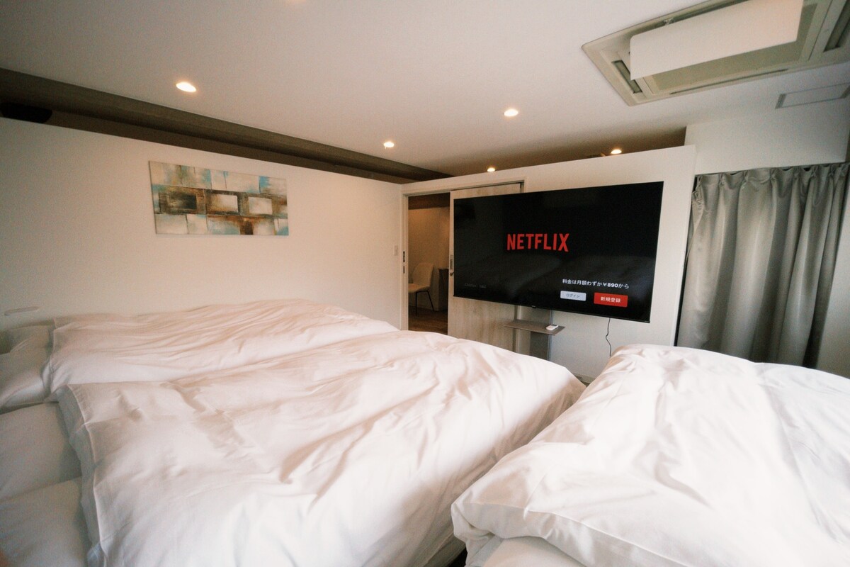 Une chambre d’hôtel avec deux lits, un téléviseur affichant le logo « NETFLIX », et un appareil de climatisation installé au plafond.