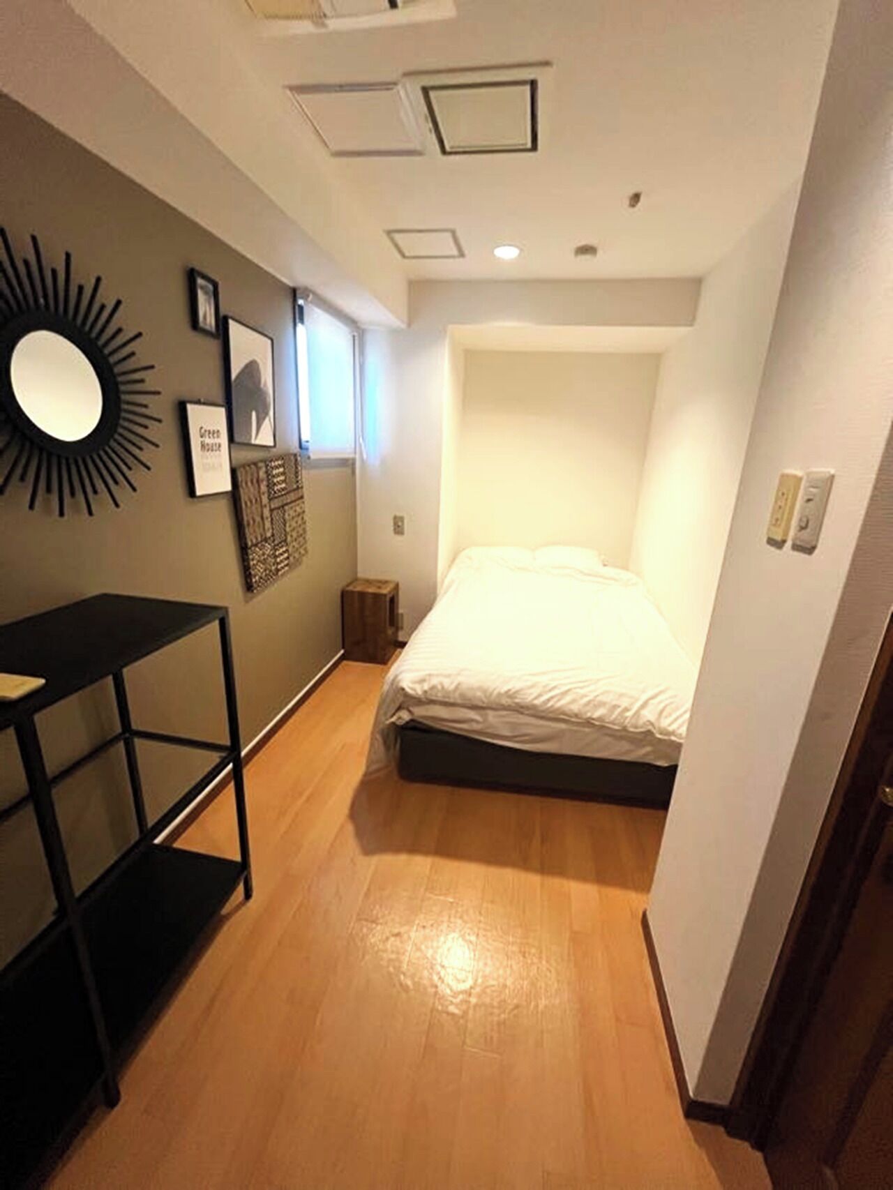 A narrow hotel room with a bed, a small table, a mirror, and framed pictures on the wall.