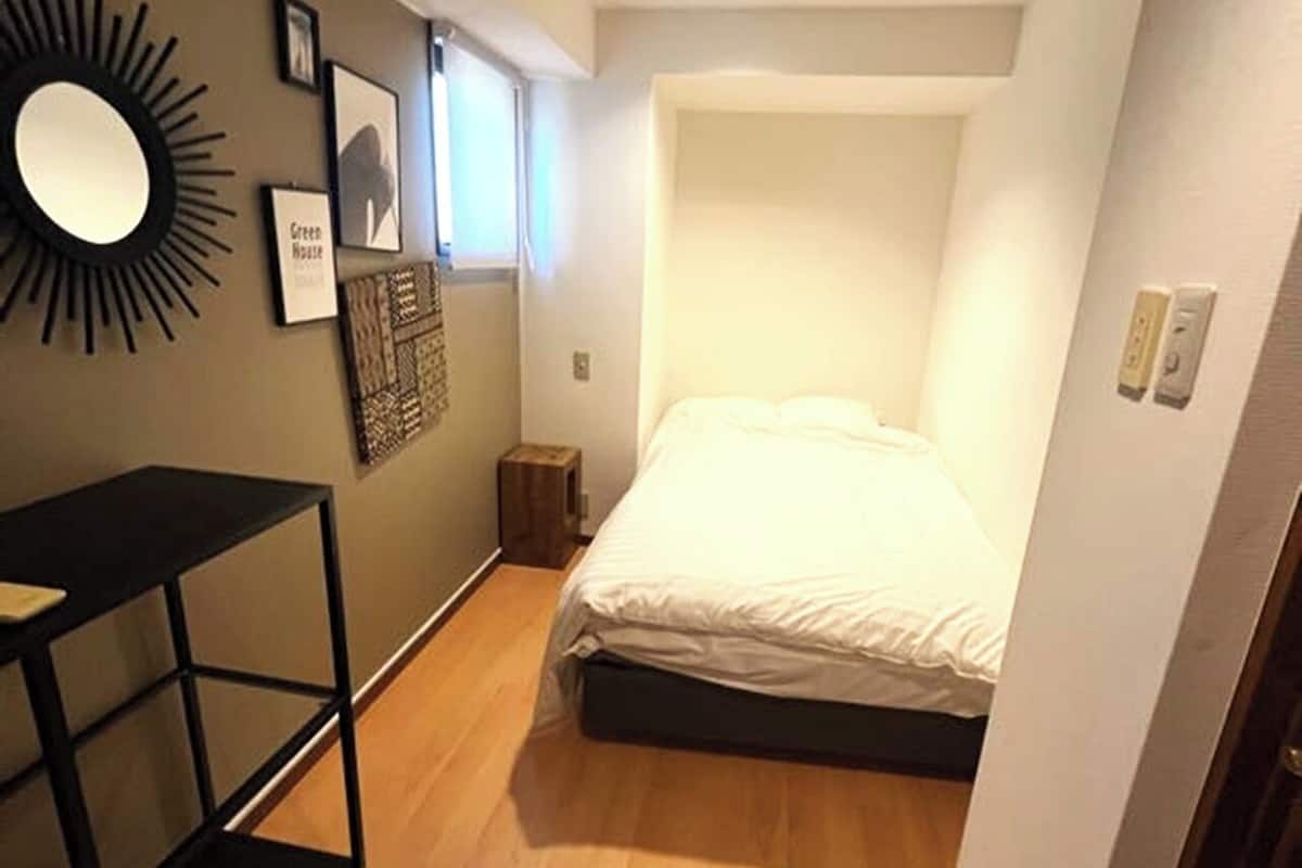 A compact bedroom with a bed, a small side table, a mirror, and framed pictures on the wall.