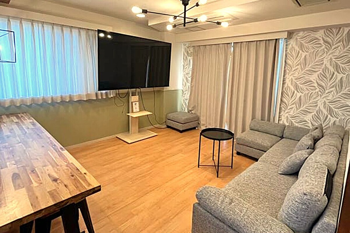 A modern living room with a wooden dining table, a grey sofa, and a flat-screen TV.