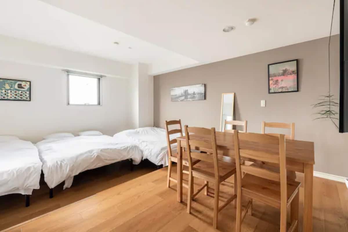 A room with a wooden dining table and chairs, a bed with white bedding, and two windows.
