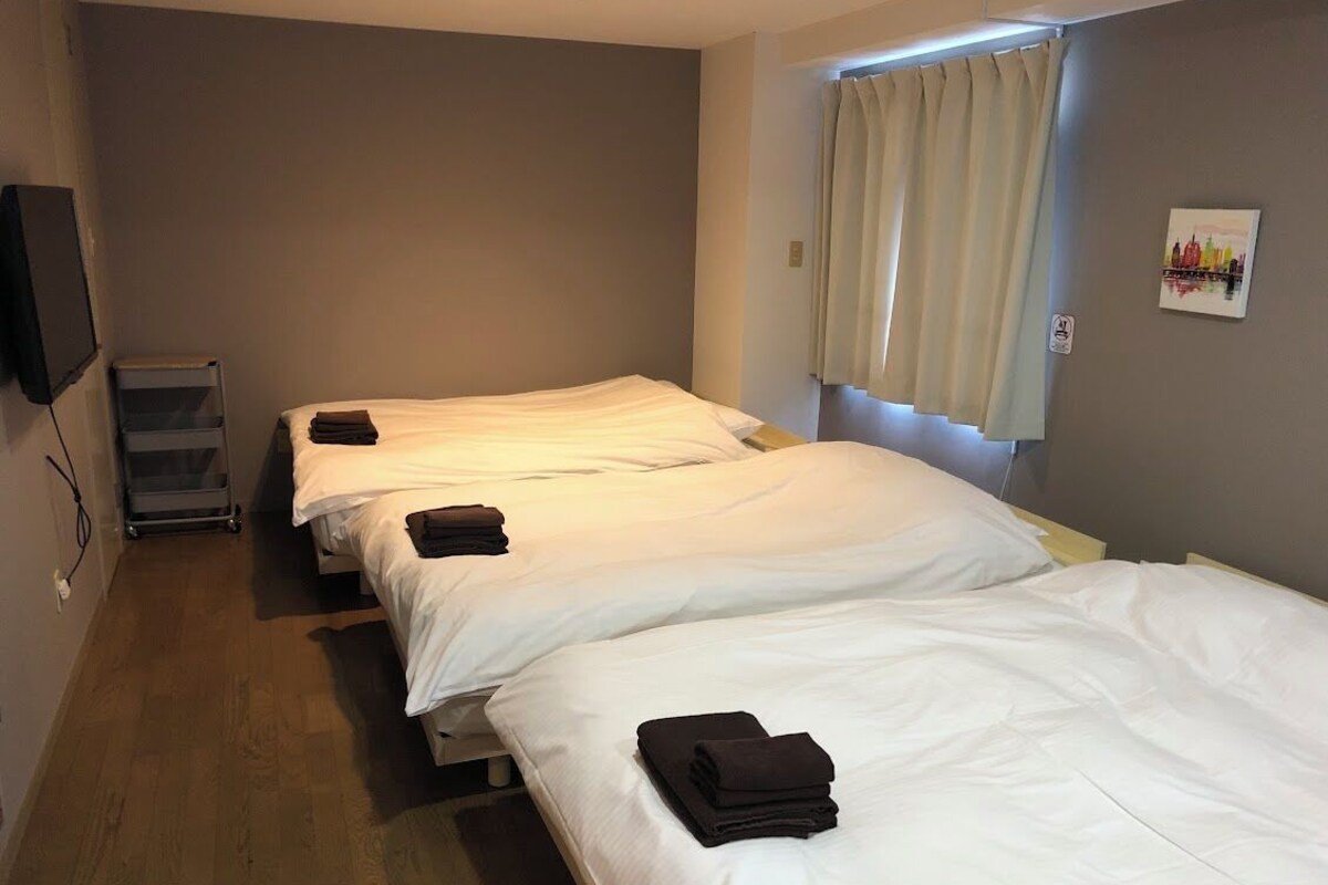 Une chambre d’hôtel avec trois lits simples, un téléviseur à écran plat fixé au mur, une fenêtre avec des rideaux et une petite œuvre d’art accrochée au mur.