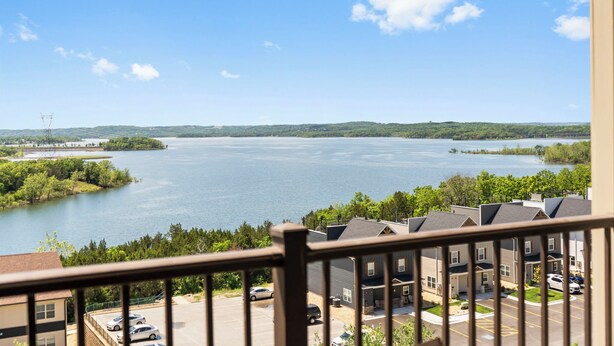Une vue sur un lac, prise depuis un balcon doté d’une balustrade, entouré de verdure et bordé d’une rangée de maisons de ville.