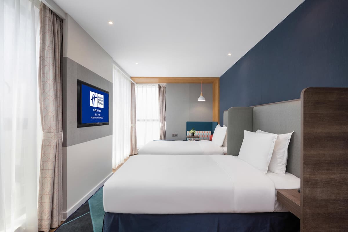 A hotel room with two beds, a TV on the wall, and a view of the city.
