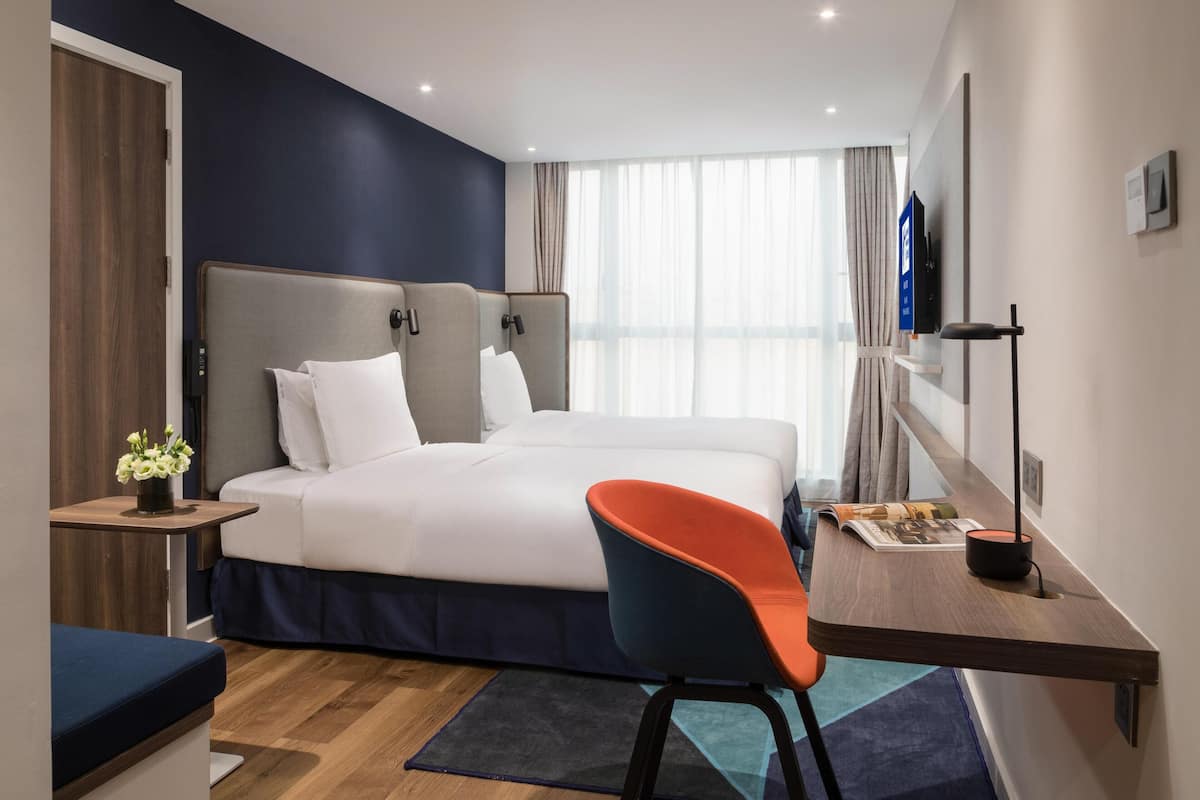 A modern hotel room with a large bed, a desk with a chair, a TV, and a window with curtains.