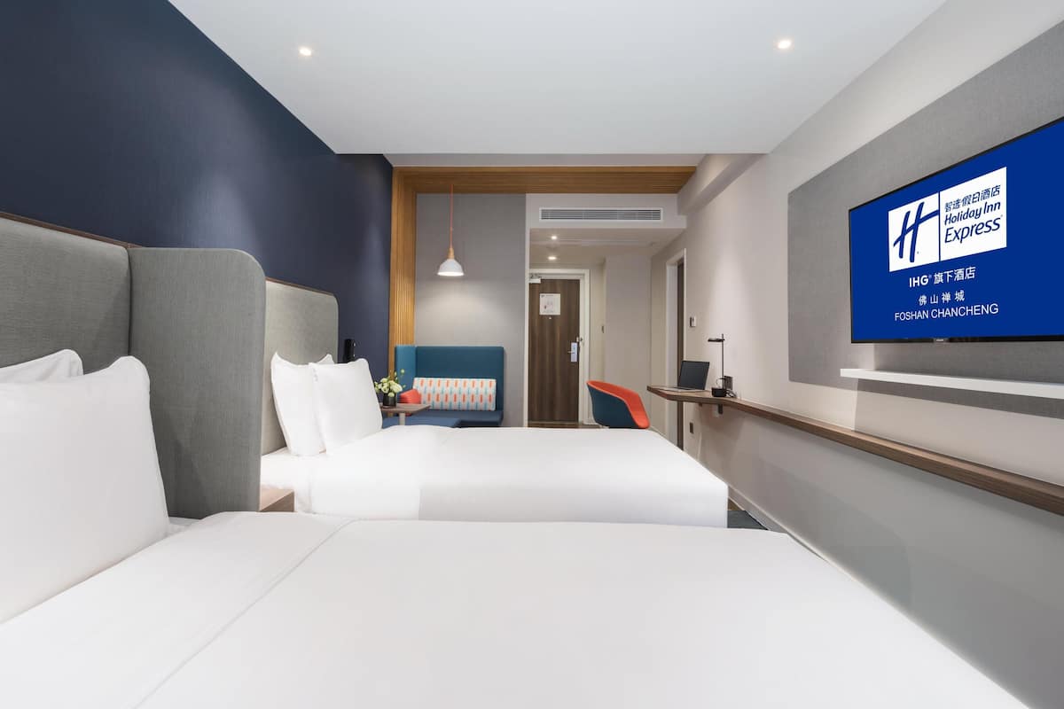 A modern hotel room with a large bed, a desk, and a television displaying the hotel name.