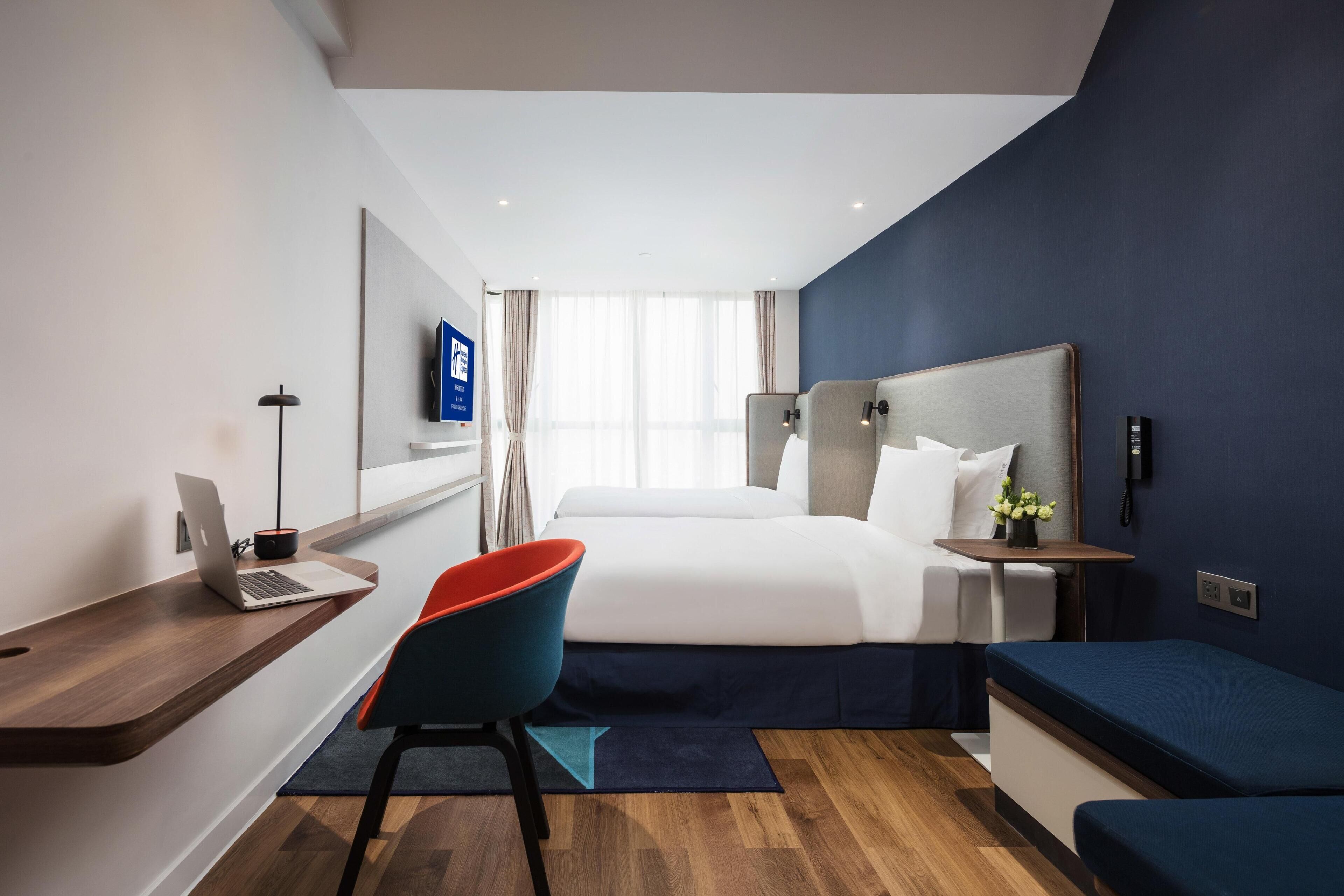A modern hotel room with a bed, desk, chair, and a small table with a vase.