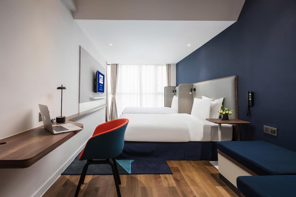 A modern hotel room with a bed, desk, chair, and a small table with a vase.