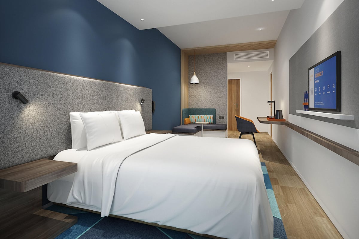 A modern hotel room with a large bed, a desk, a chair, and a TV mounted on the wall.