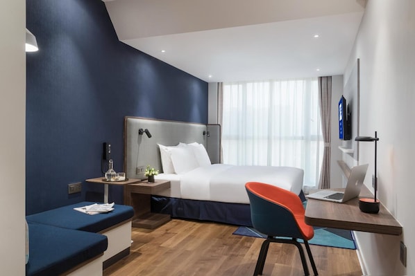 A modern hotel room with a bed, a desk with a laptop, a chair, and a bedside table with a lamp.