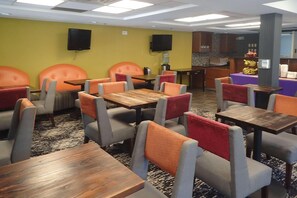 A dining area with multiple tables and chairs, a kitchen area in the background, and a yellow wall with two speakers mounted on it. - Clarion Inn Atlantic City - Beach and Boardwalk (Atlantic City)