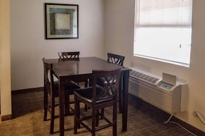 A dining area with a table and chairs, a framed picture on the wall, and a window with blinds. - Clarion Inn Atlantic City - Beach and Boardwalk (Atlantic City)