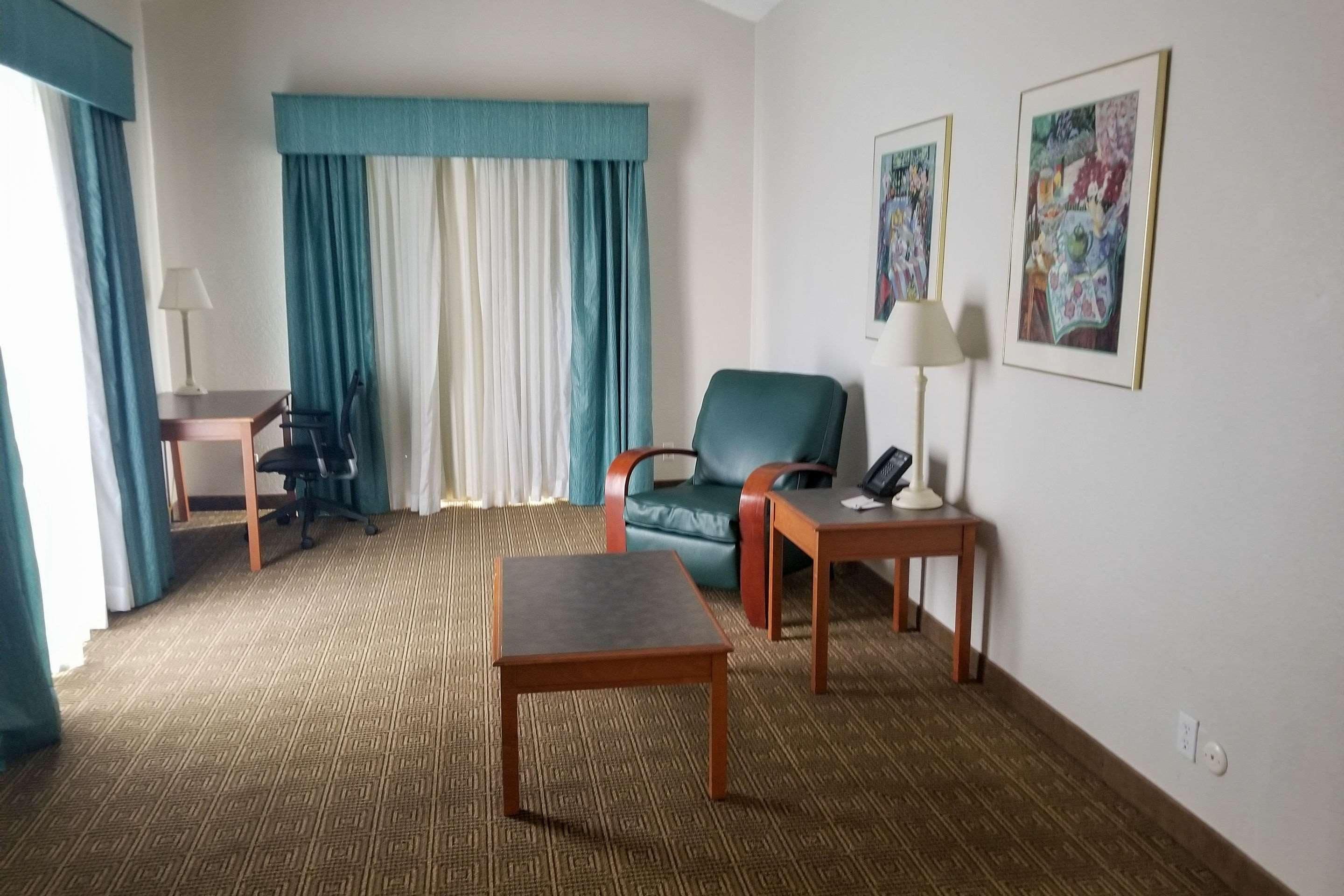 A hotel room with a bed, a desk, a chair, a table, a lamp, and two paintings on the wall.