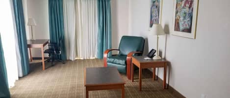 A hotel room with a bed, a desk, a chair, a table, a lamp, and two paintings on the wall.