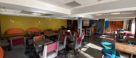 A spacious dining area with multiple tables and chairs, a kitchen in the background, and a yellow accent wall.