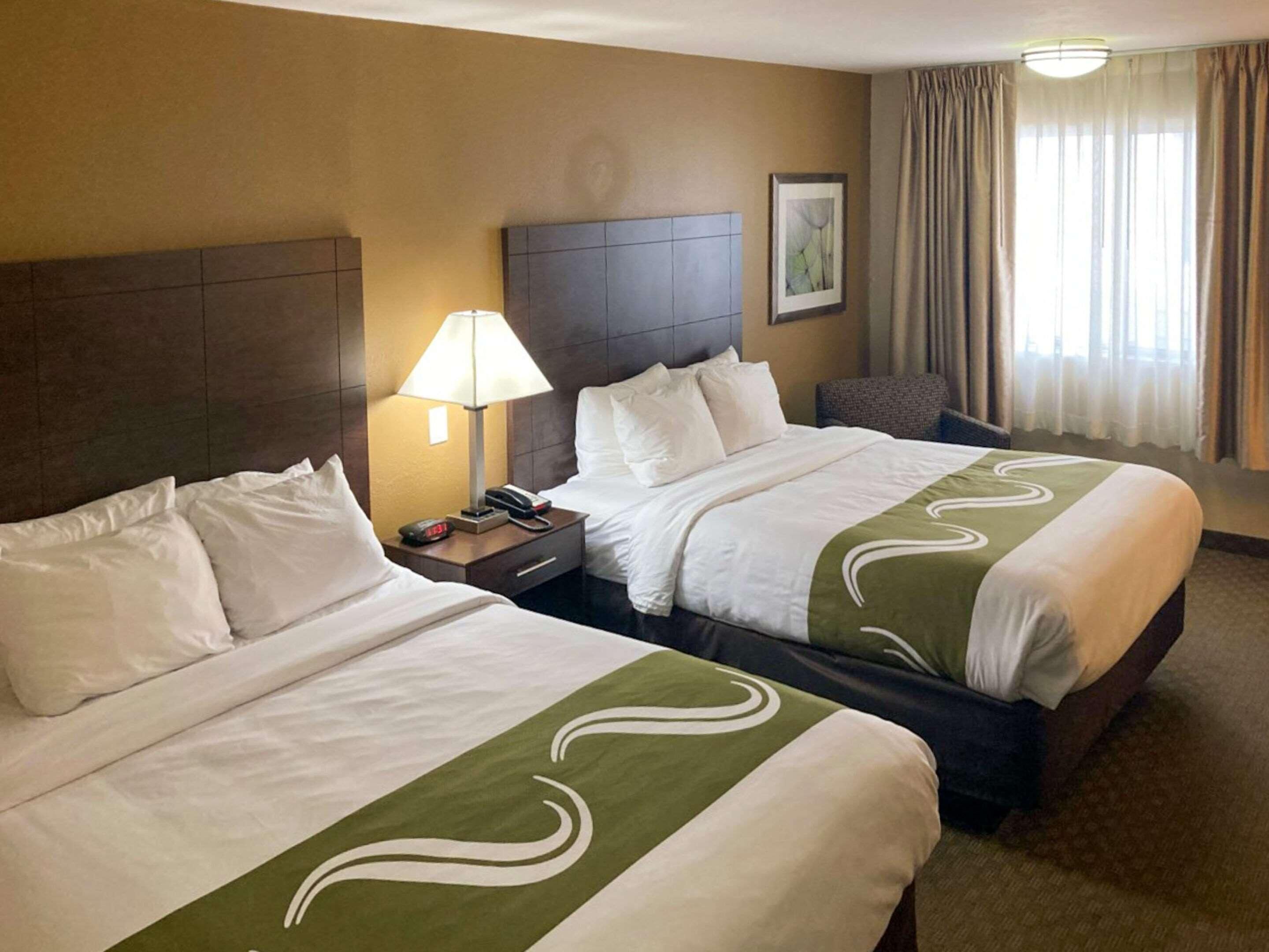 A hotel room with two beds, a nightstand with a lamp, a chair, and a window with curtains.