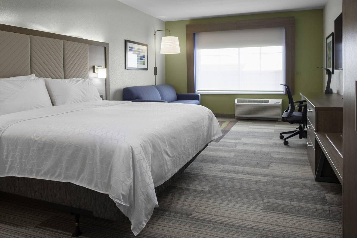 A hotel room with a large bed, a desk, a chair, a window with blinds, and a wall-mounted air conditioner.