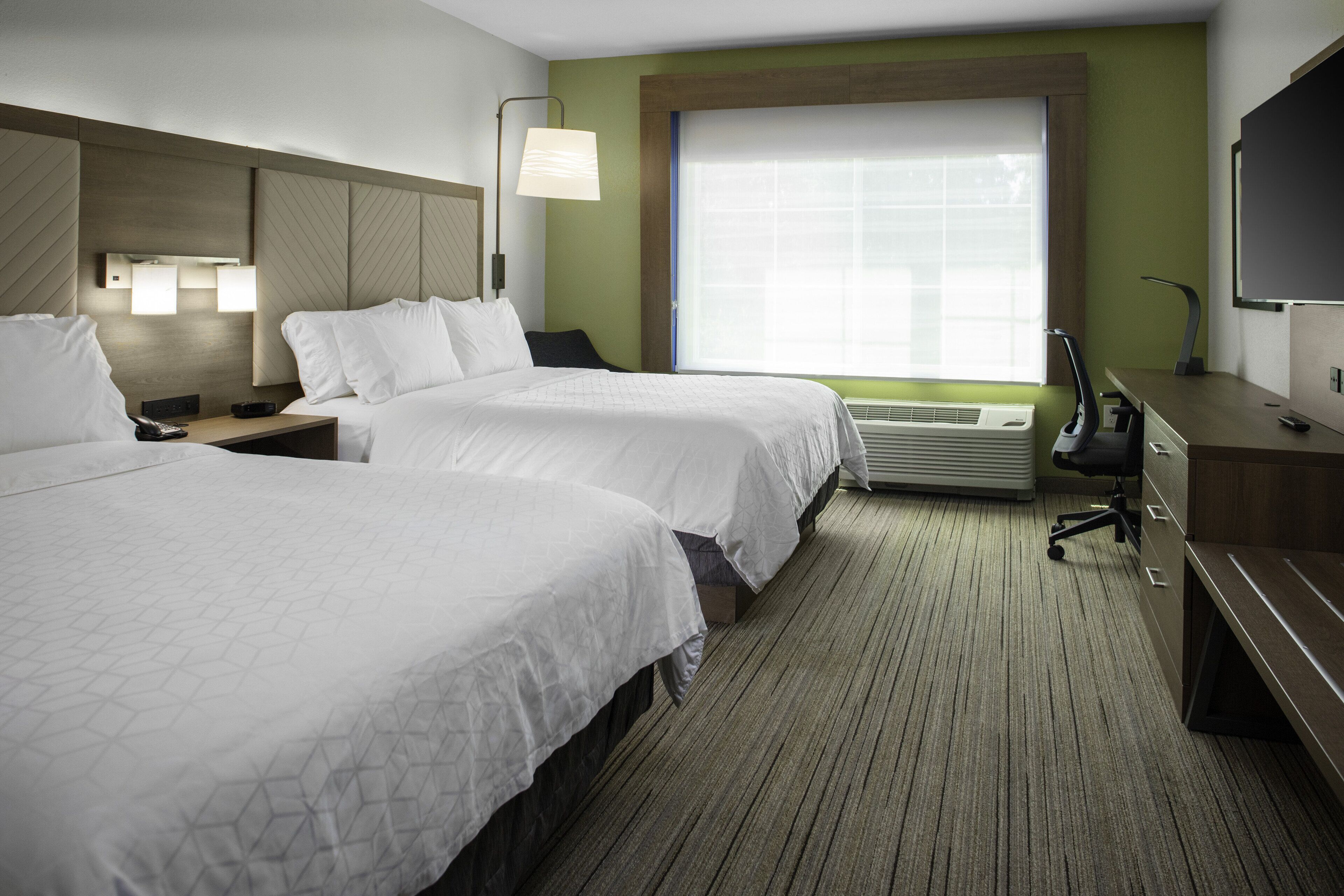 A hotel room with two beds, a desk, a chair, a TV, and a large window with blinds.