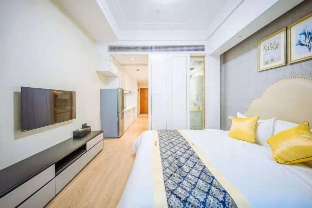 A modern hotel room with a bed, bedside table, TV, and a view into the hallway.