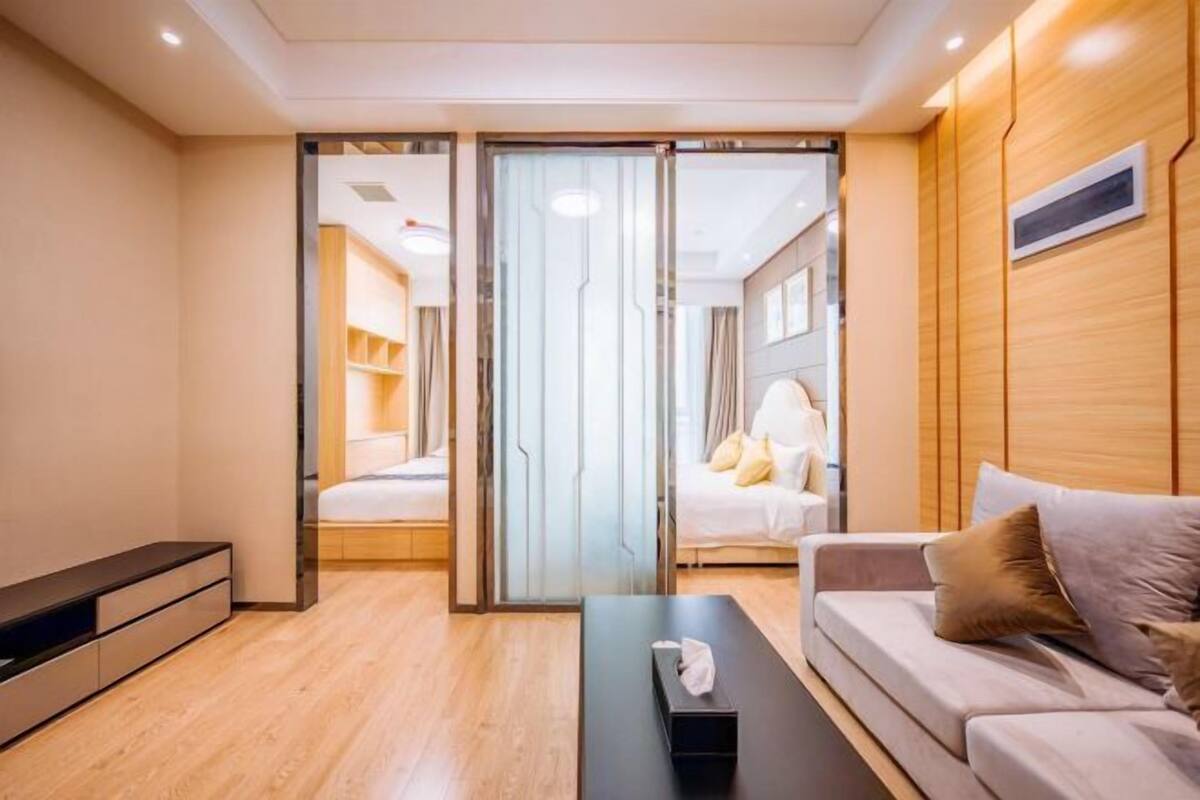 A modern hotel room with a sofa, a bedside table, a glass partition, and a bed with a white headboard.