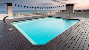 Una piscina rectangular con cubierta de madera, rodeada por una barandilla, con vista a una playa y edificios a lo lejos.