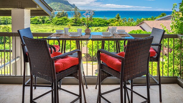 A balcony with wicker chairs and a table set for four, overlooking a lush landscape and the ocean.