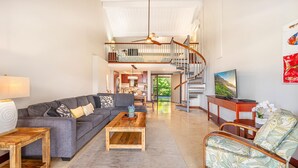 A spacious living room with a grey sofa, wooden coffee table, and a patterned armchair. A staircase leads to an upper level with a balcony.