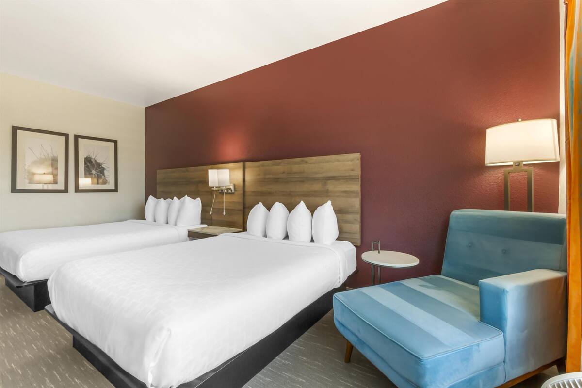 A hotel room with two beds, a blue armchair, a round table, and a wall-mounted lamp.