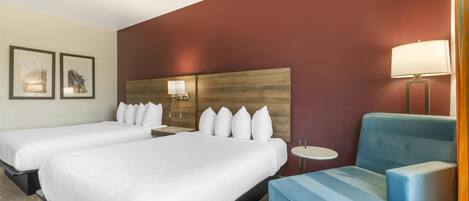 Habitación de hotel con dos camas, un sillón azul, una mesa redonda y una lámpara de pared.