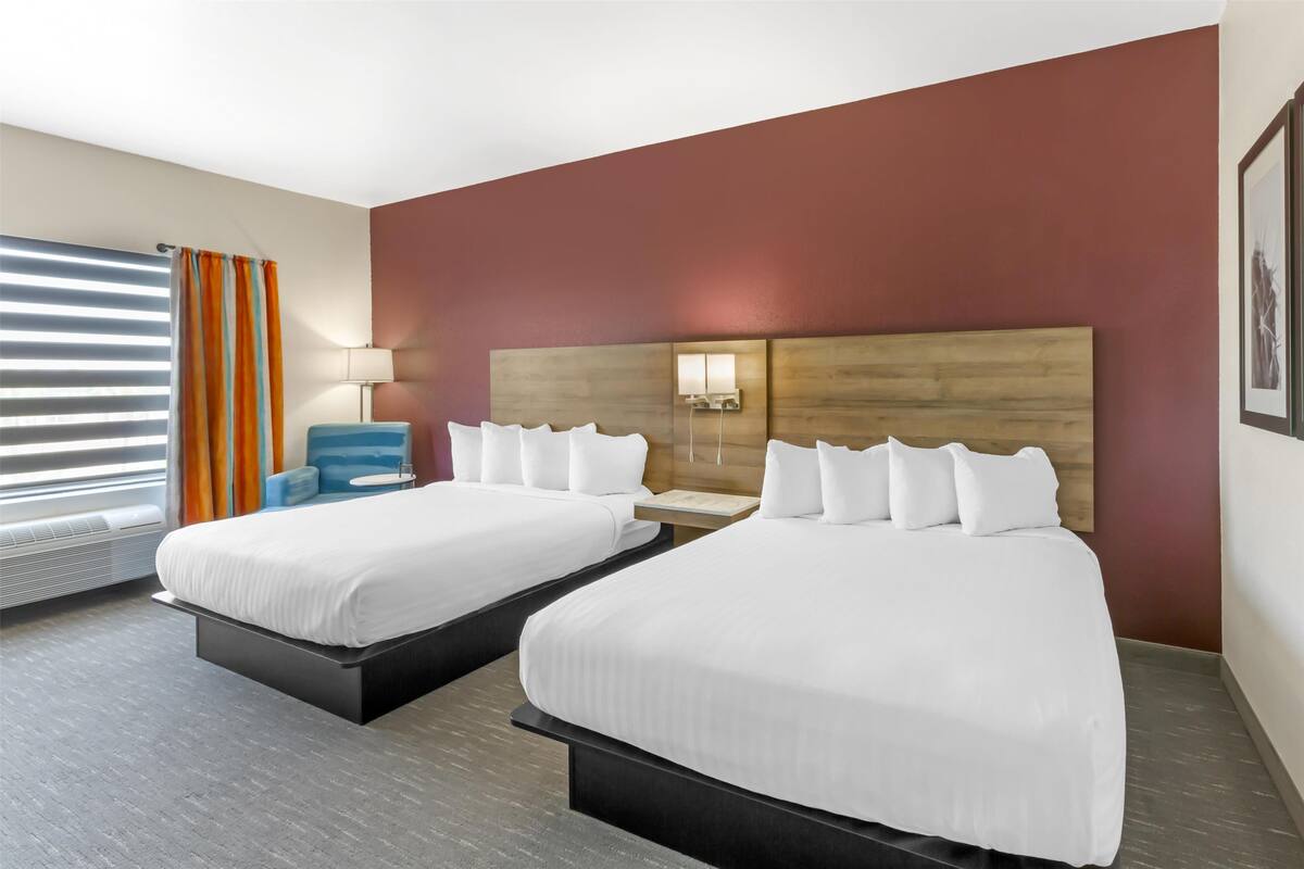 A hotel room with two beds, a nightstand, a lamp, and a window with blinds.