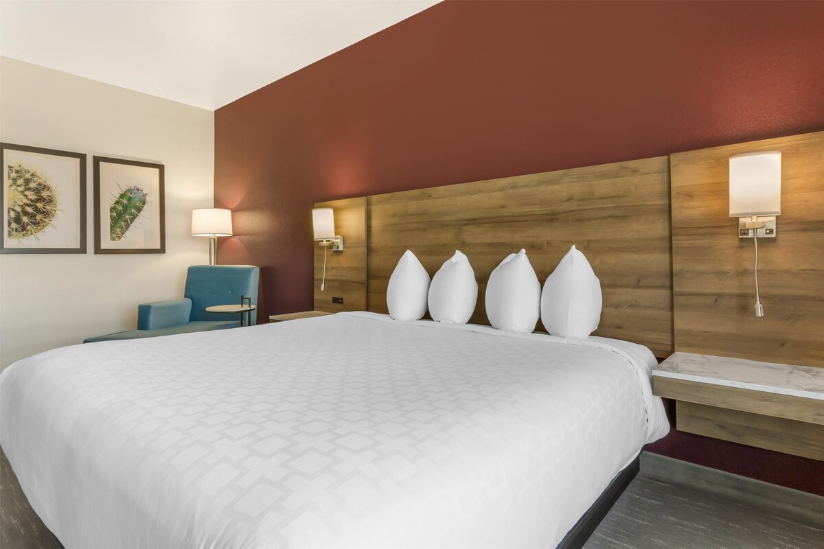 A hotel room with a large bed, a wooden headboard, wall-mounted lights, a small round table, and two framed cactus artworks.