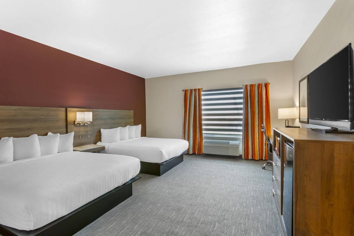 A hotel room with two beds, a TV, a desk, and a window with curtains.