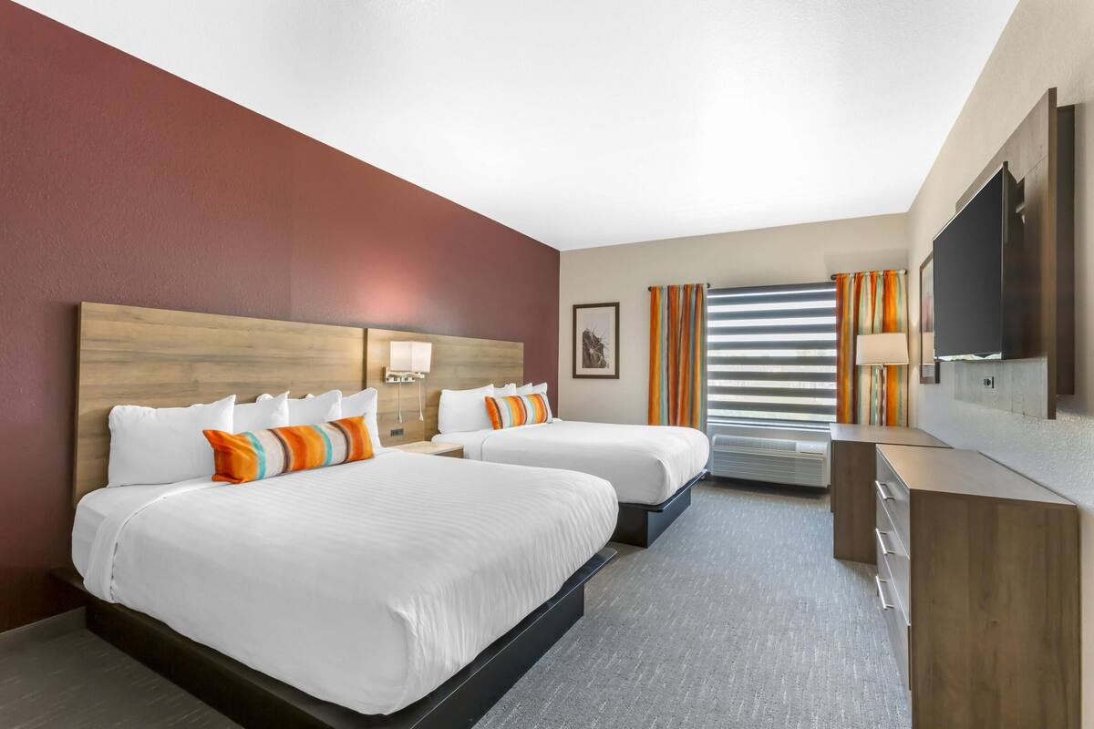 A hotel room with two beds, a flat-screen TV, a desk, and a window with blinds.