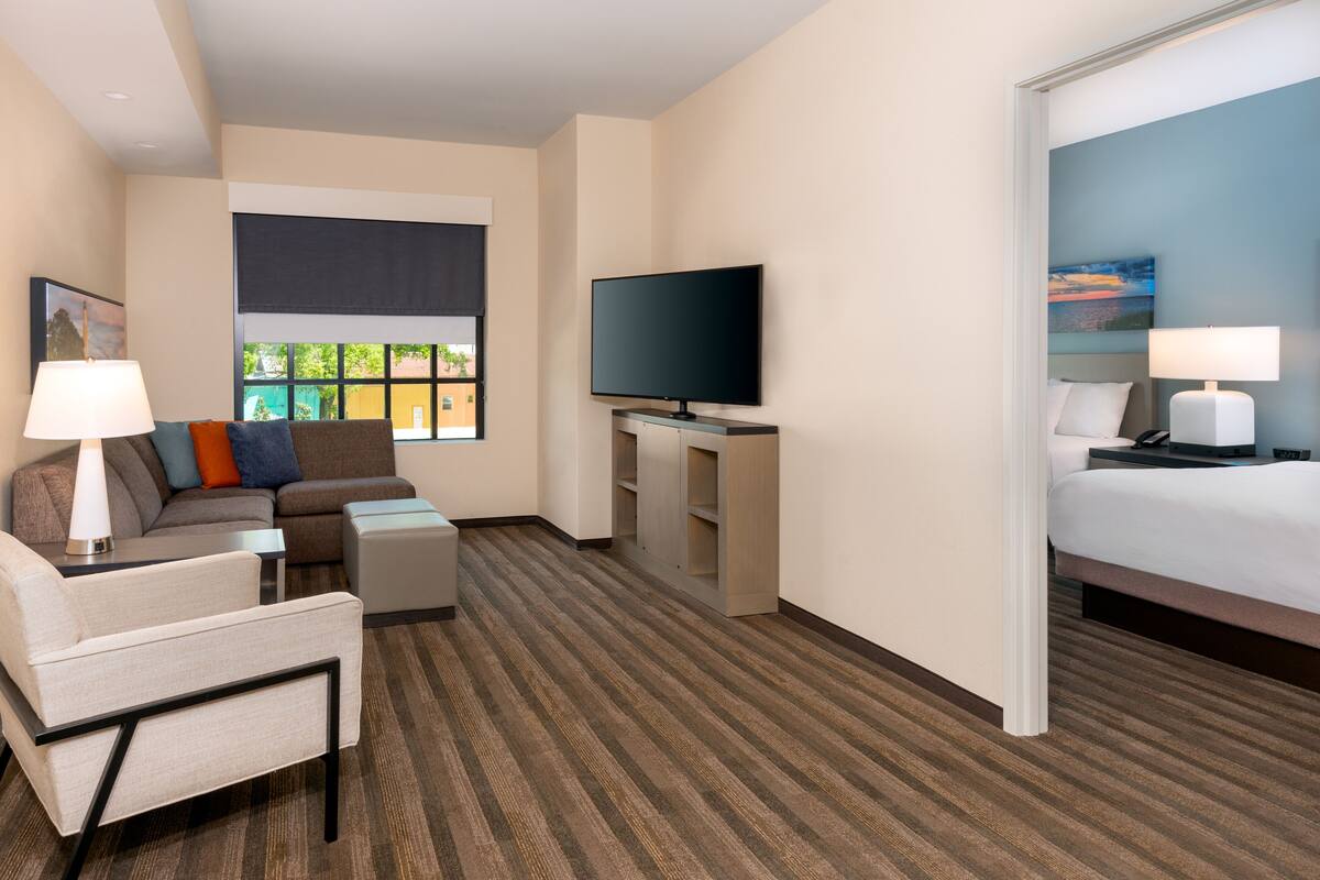 Una habitación de hotel con sofá, sillón, televisor, mesita de noche, lámpara y cama.