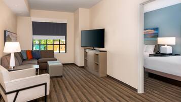 A hotel room with a sofa, armchair, TV, bedside table, lamp, and a bed.