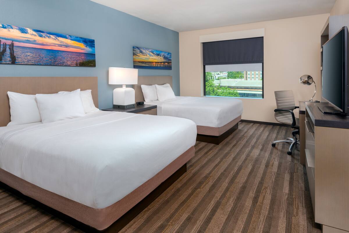 Habitación de hotel con dos camas, un escritorio con computadora, un televisor y dos cuadros en la pared.