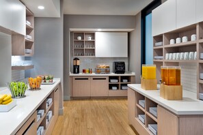 A modern kitchen with wooden cabinets, a central island with a coffee machine, and a breakfast bar with fruit and juice dispensers.