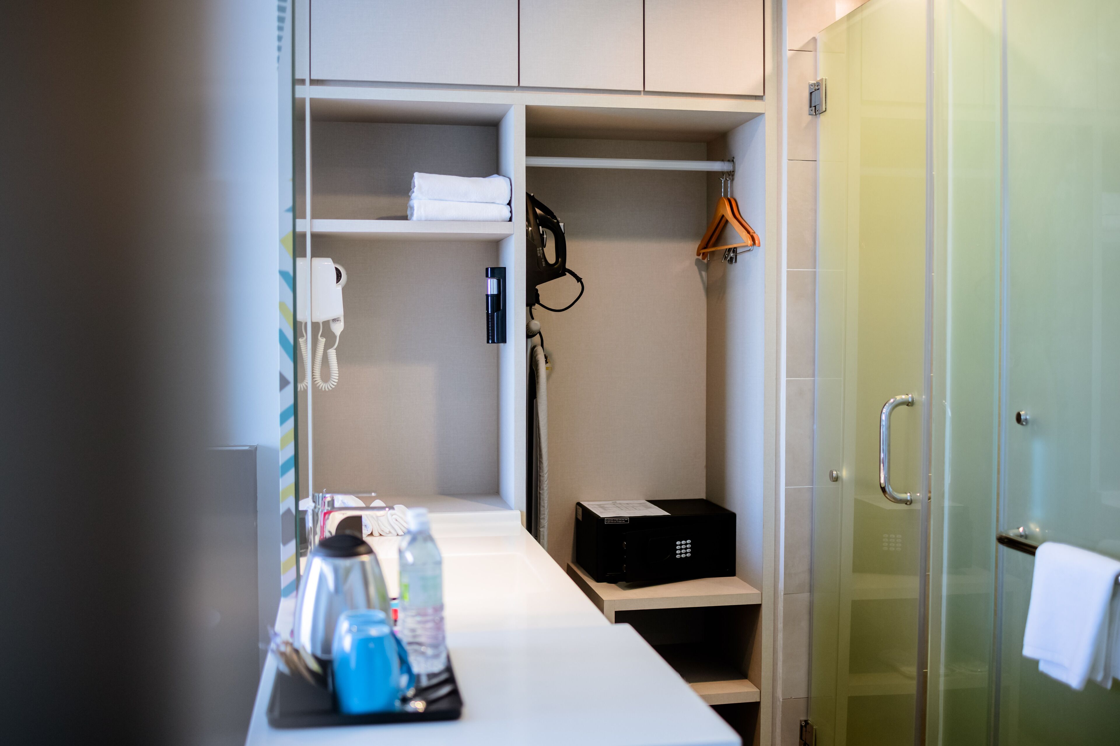 A compact hotel room with a kitchenette, a glass shower enclosure, and a neatly organized storage area with towels and toiletries.