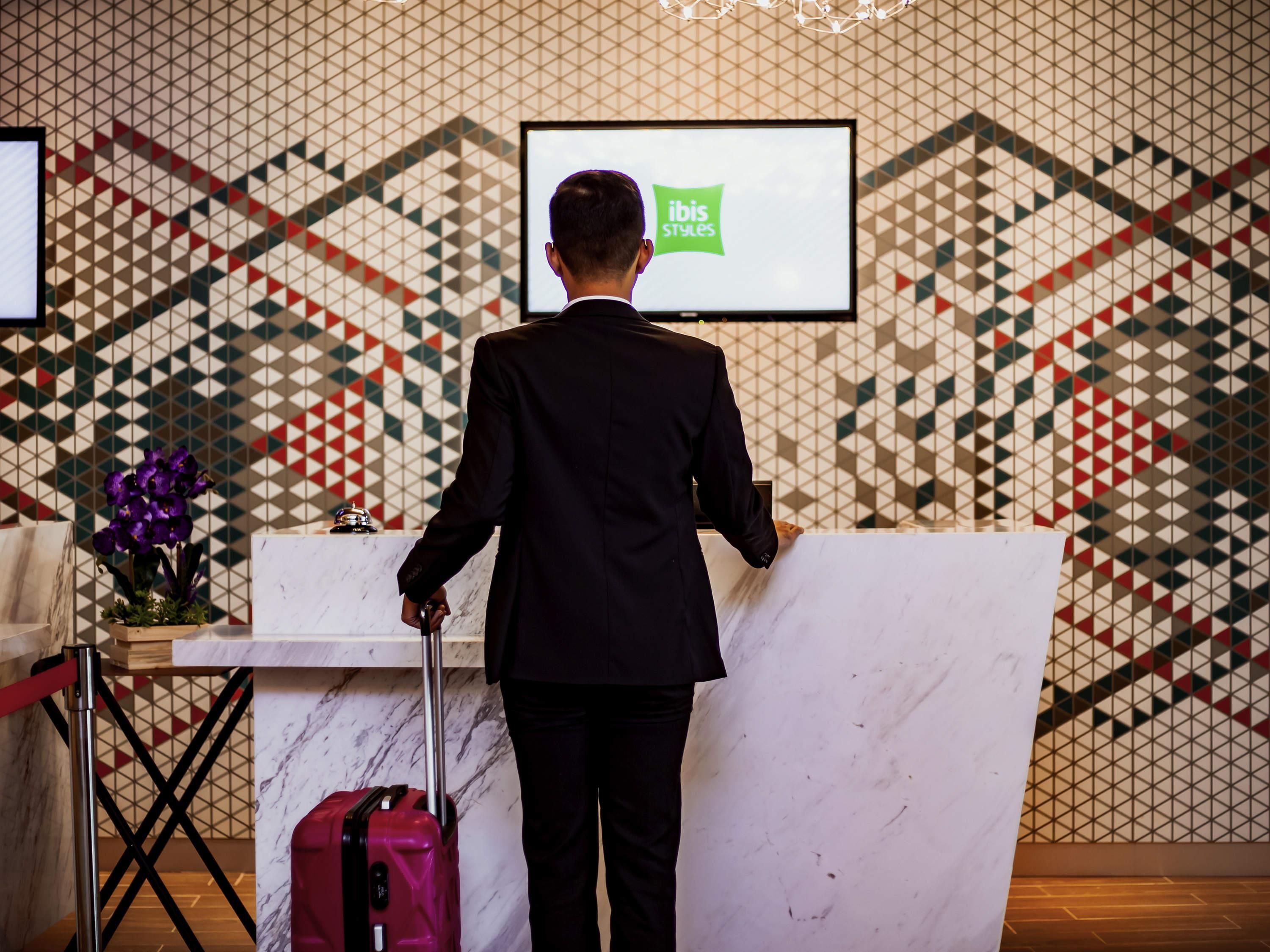 A man in a suit standing at a reception desk with a laptop, a red suitcase, and a potted plant.