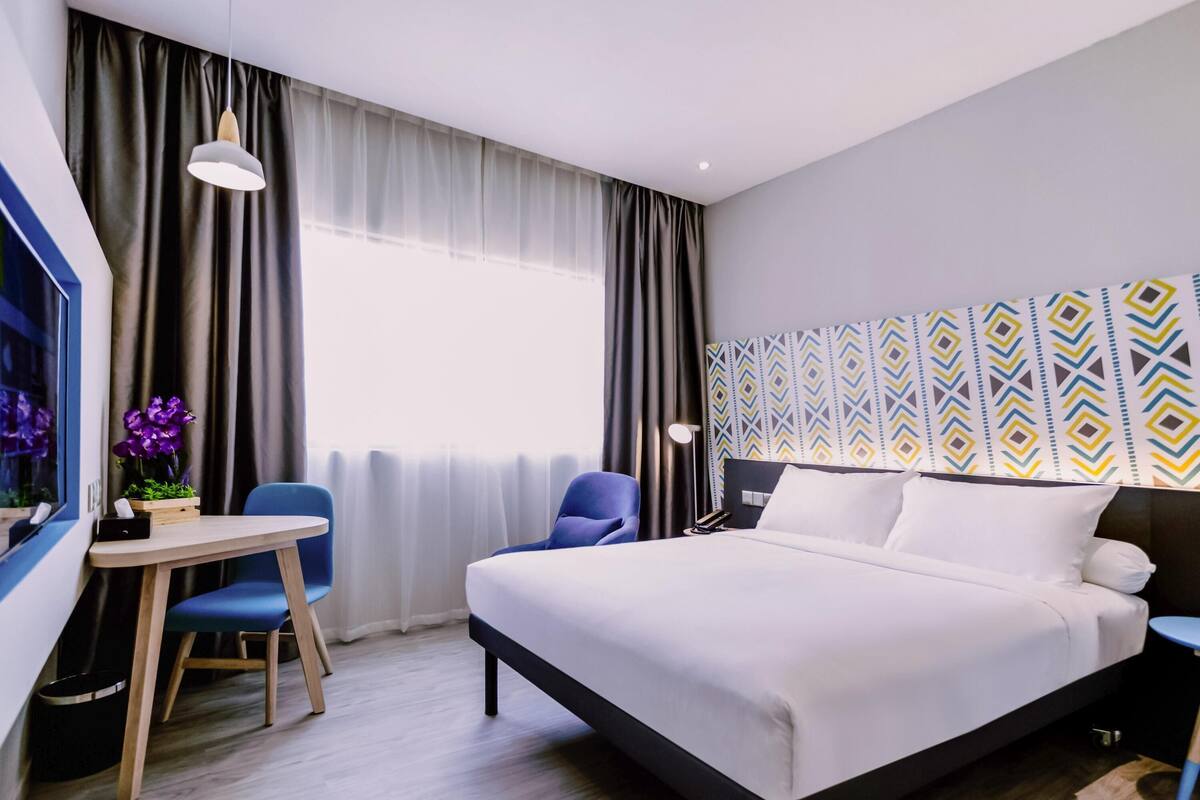 Una habitación de hotel con cama, escritorio, silla, mesita de noche, lámpara y ventana con cortinas.