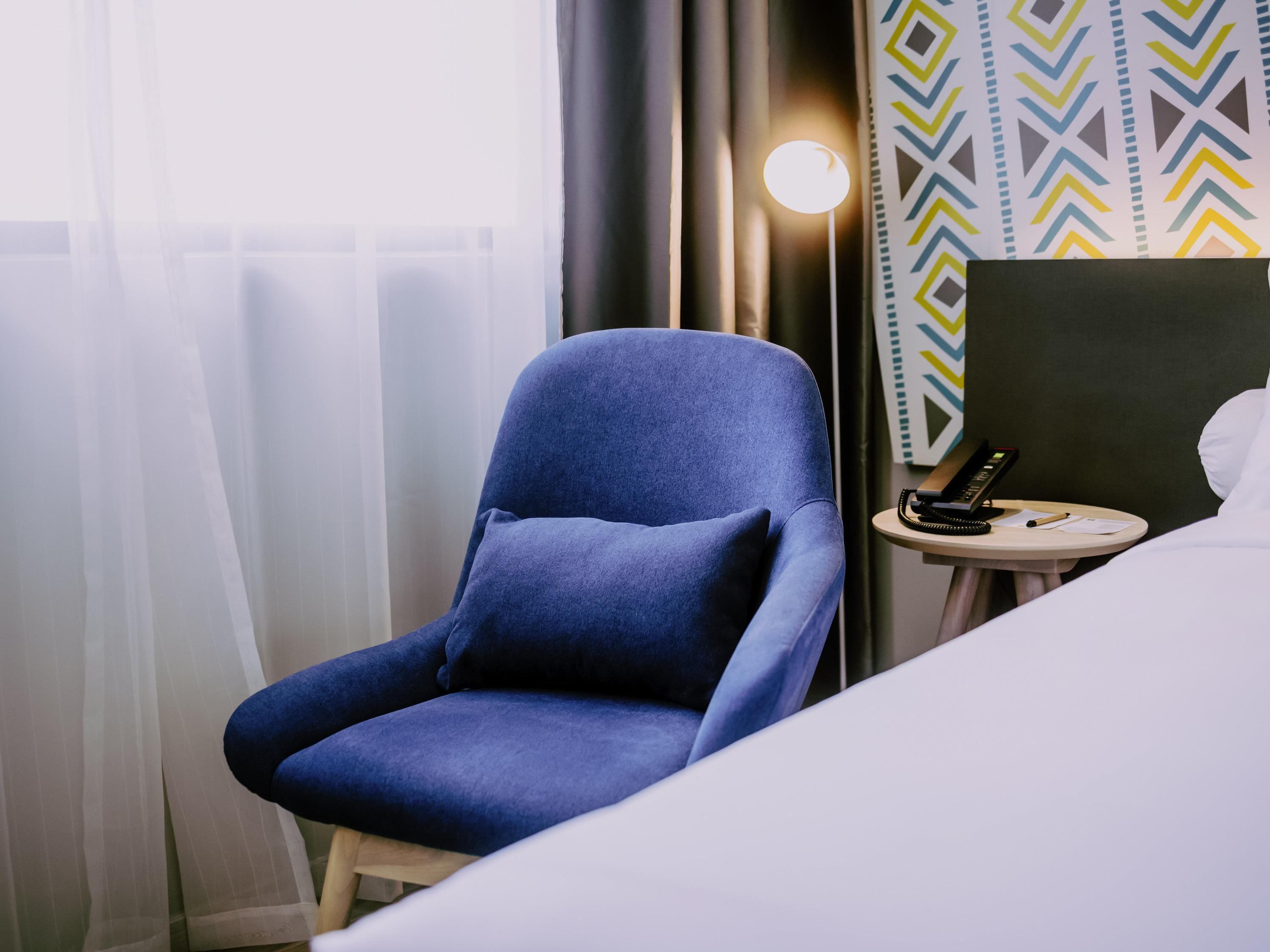 A hotel room with a blue armchair, a bedside table with a phone and papers, and a bed with white linens.