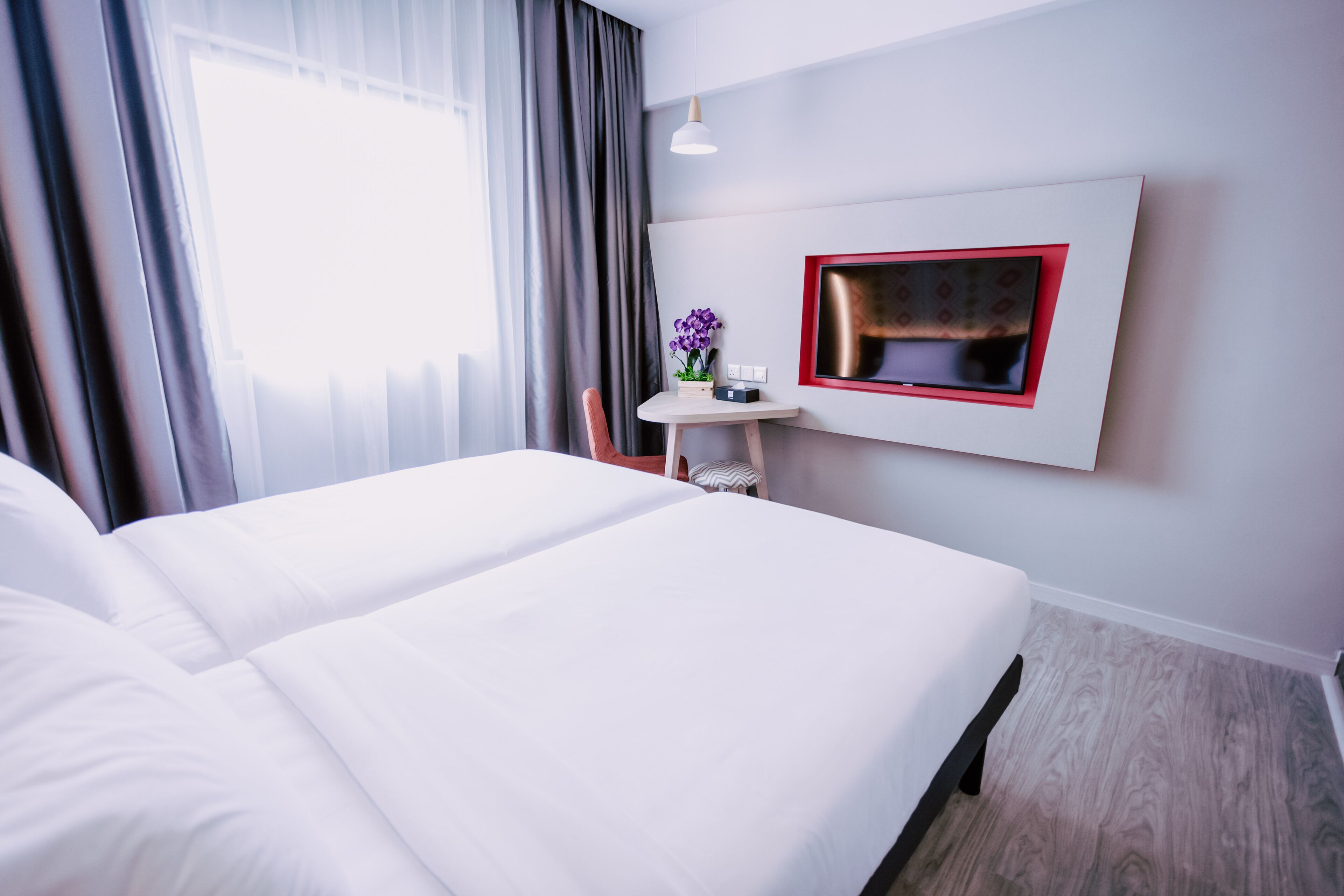 A hotel room with a large bed, a small table with a vase of flowers, a mounted TV, and a window with curtains.