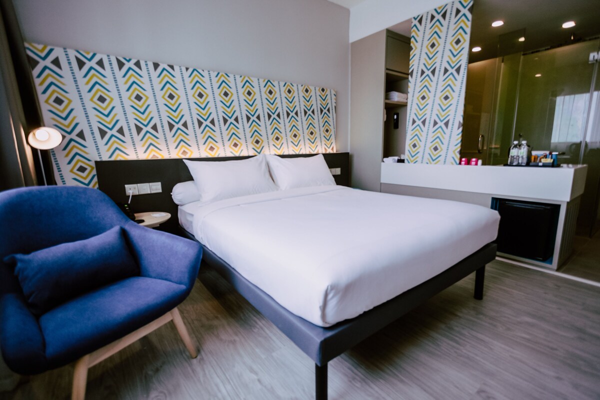 A modern hotel room with a large bed, a blue armchair, a bedside table, and a mirror.