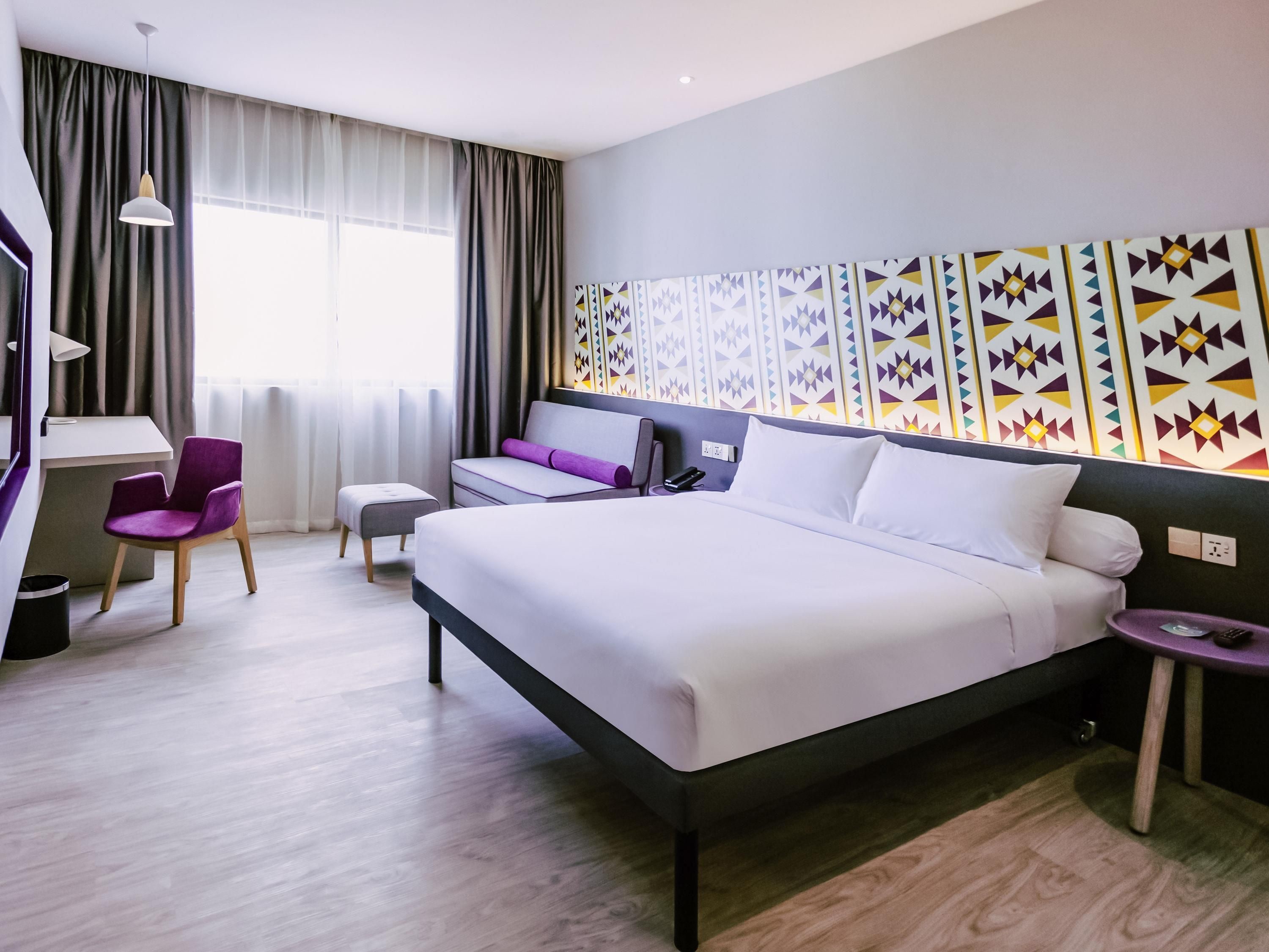A modern hotel room with a large bed, a desk, a chair, and a wall with patterned decor.