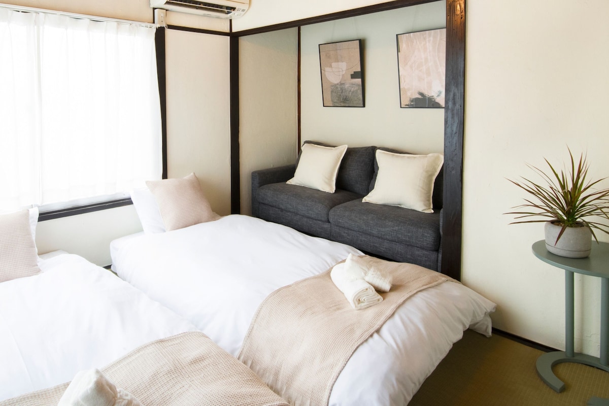 A hotel room with two beds, a grey sofa, a small table with a plant, and artwork on the wall.