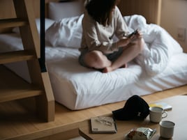 Iemand zit op een bed met een notitieboek, een bril en een mok op een houten tafel.